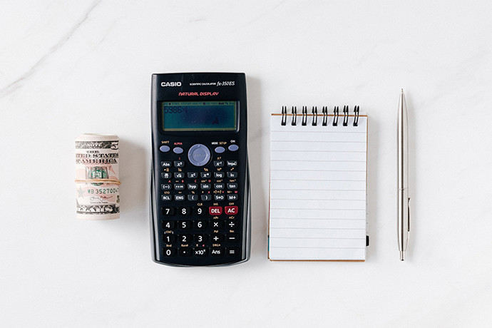 Roll of money, calculator, notepad, and a pen located on white table